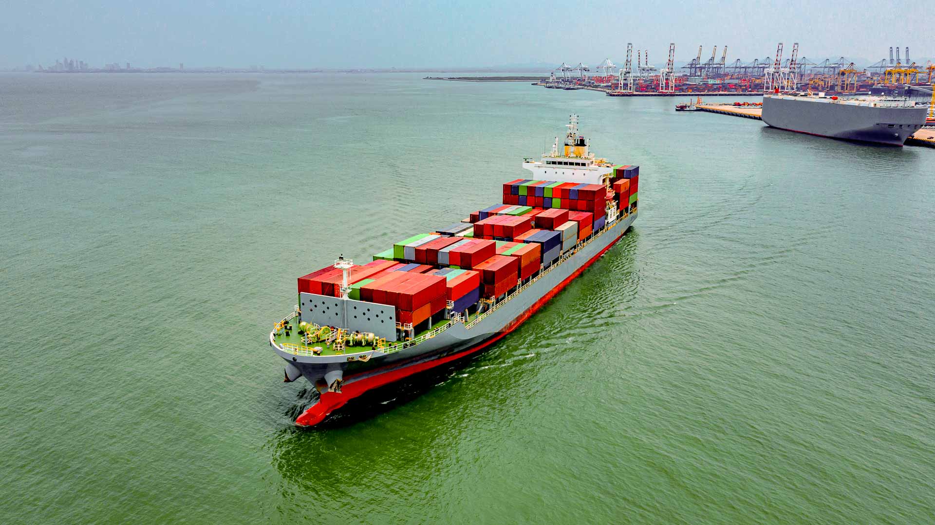 Container ship sailing into a busy port terminal lined with cranes and vessels, carrying multicolored cargo units—symbolizing the global flow of goods and the need for smart, scalable port coordination.