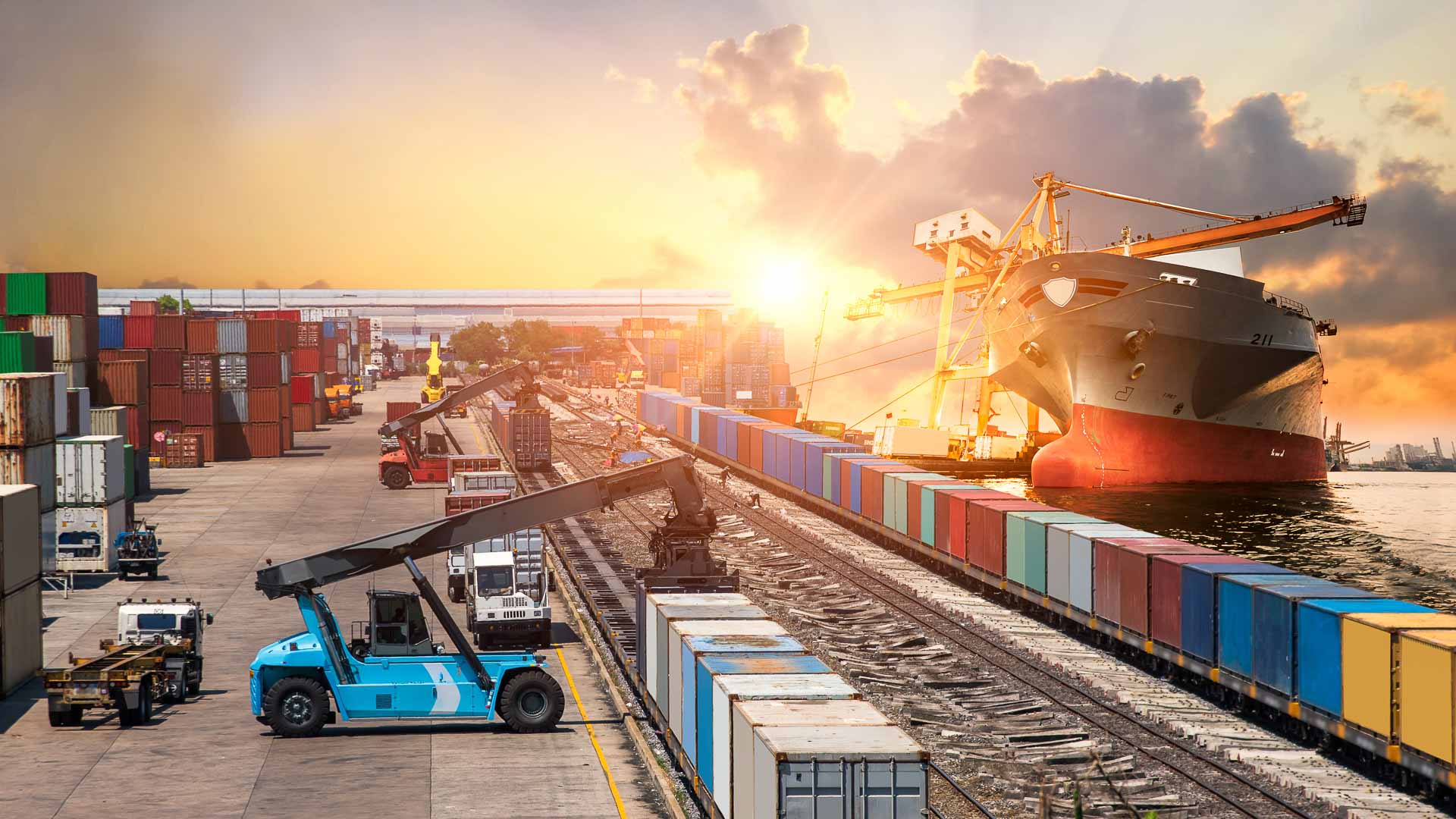 A cargo ship docked at a bustling port terminal during sunrise, with colorful shipping containers lined up alongside railway tracks, highlighting the complexity and scale of global logistics.