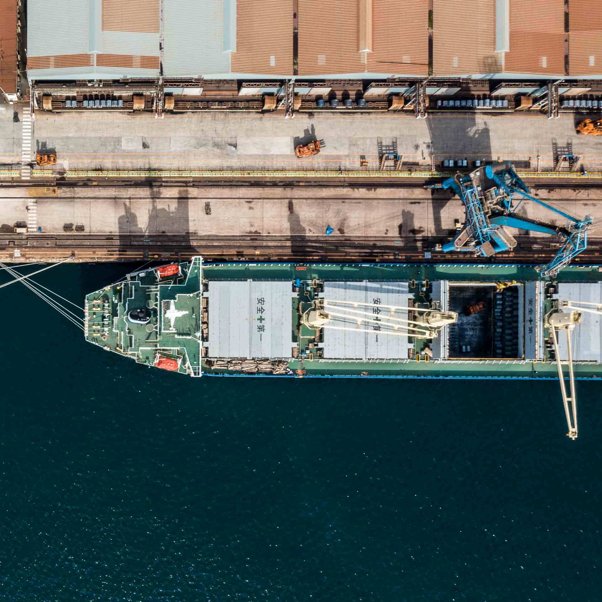 Top-down view of a bulk cargo ship docked at a port terminal, with cranes positioned for loading or unloading—illustrating operational precision and infrastructure alignment in port logistics.