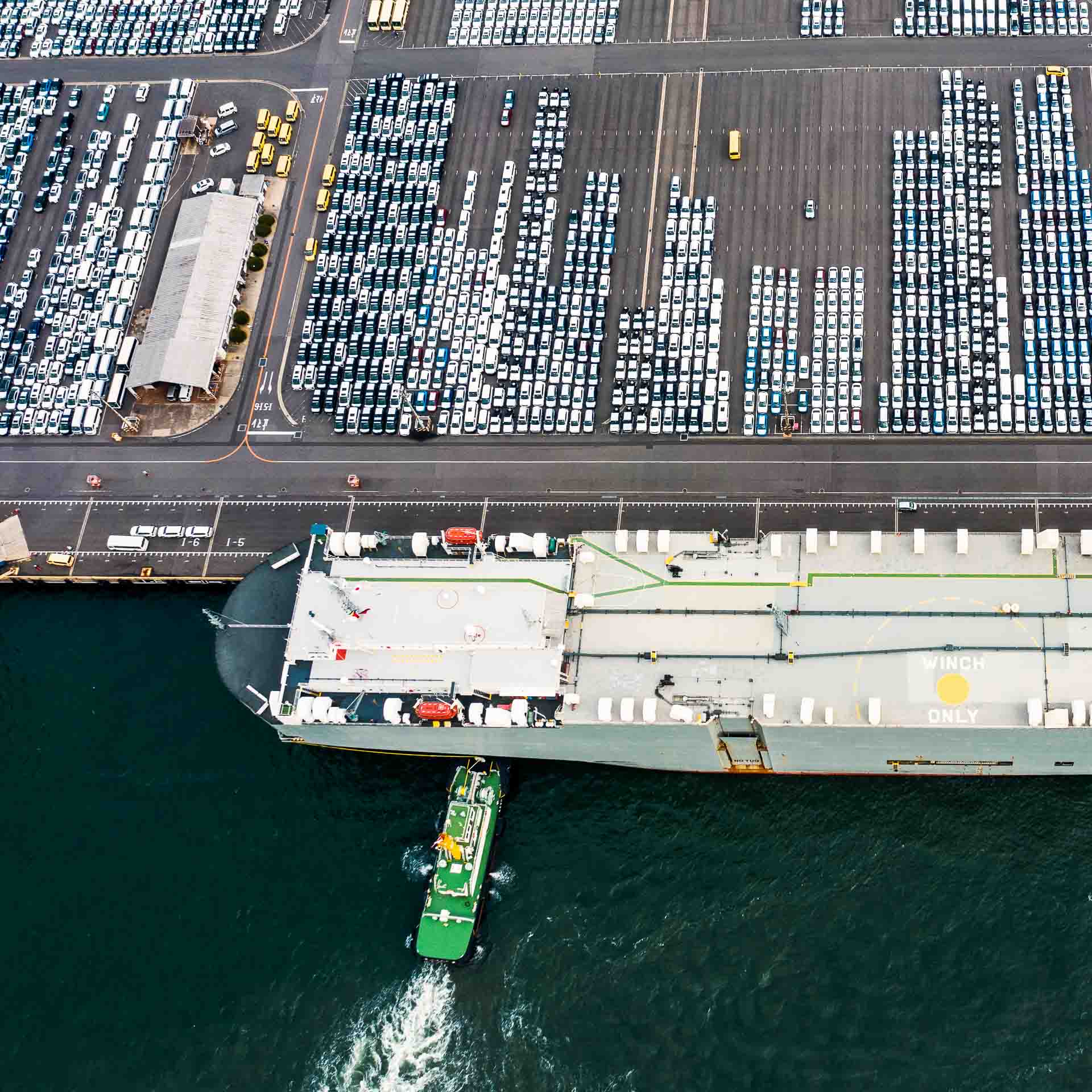 Aerial view of a roll-on/roll-off cargo ship docked at a port terminal with rows of parked vehicles—highlighting the scale, structure, and logistics behind vehicle import and export operations.